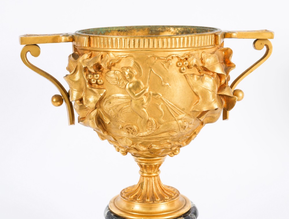 A pair of nineteenth-century French gilt bronze classical urns featuring relief carvings and scroll handles, mounted on dark green marble bases. View XVIII