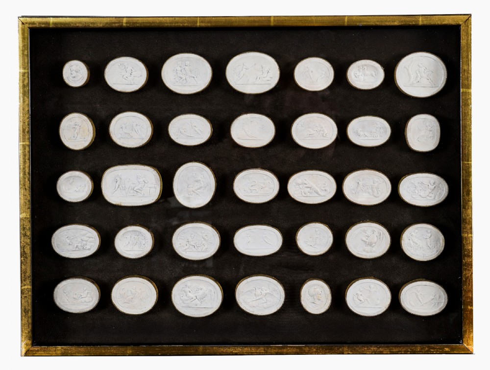 A framed display of thirty-five off-white plaster intaglio impressions featuring classical relief figures mounted on a dark background within a gilt frame.