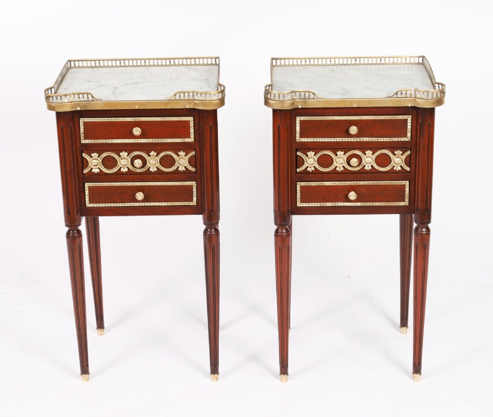 A pair of 19th-century mahogany French Empire bedside cabinets featuring white marble tops with pierced brass galleries, gilt bronze mounts, and slender fluted legs.