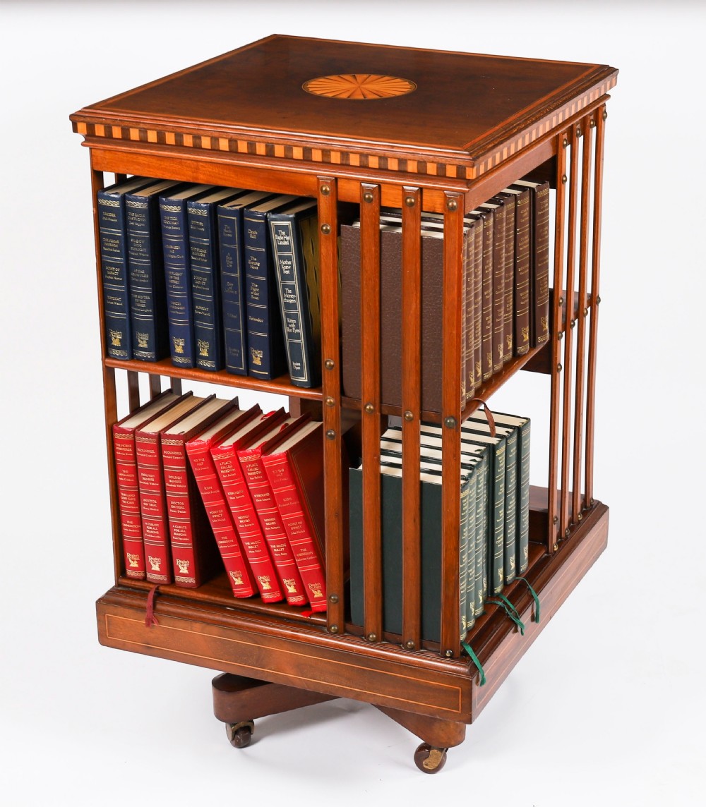 An Edwardian flame mahogany revolving bookcase featuring two tiers, vertical slatted sides with brass studs, satinwood inlay, and castors.