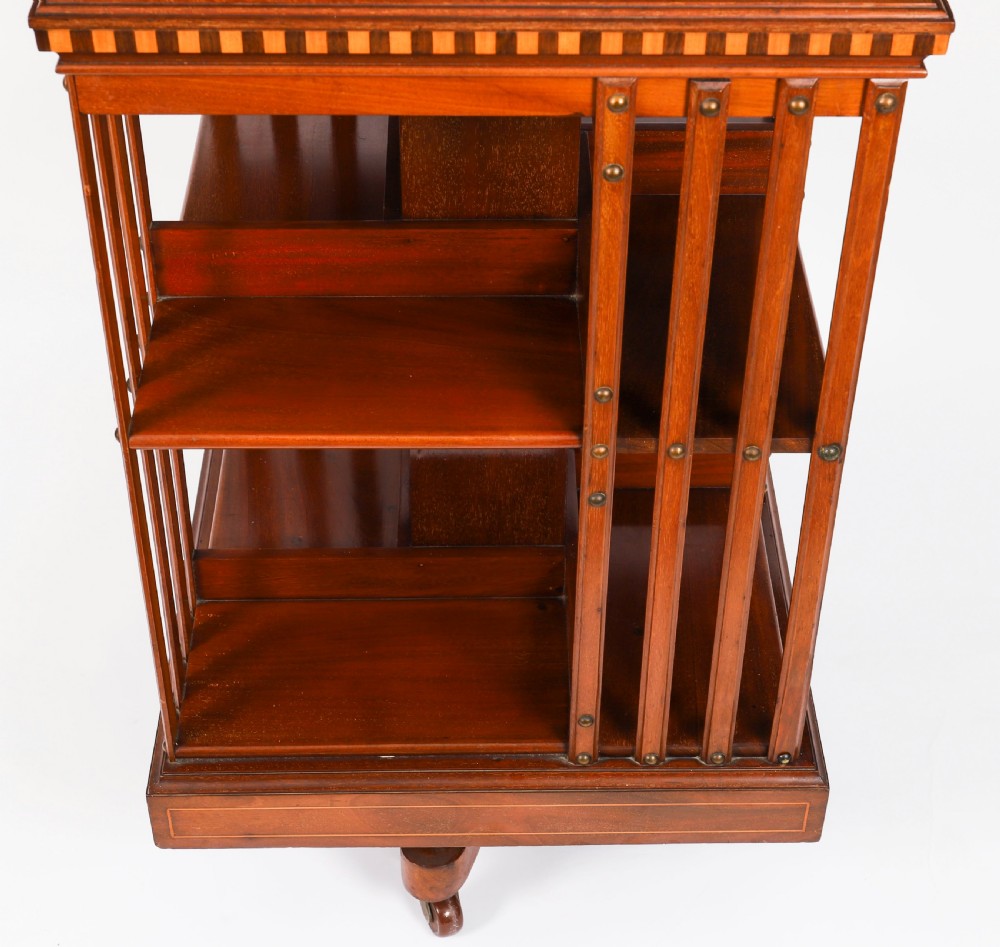 An Edwardian flame mahogany revolving bookcase featuring two tiers, vertical slatted sides with brass studs, satinwood inlay, and castors. View X