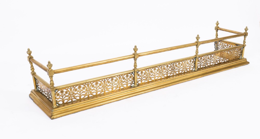 A 19th-century Victorian polished brass fireplace fender featuring a low pierced floral base band topped with a tubular rail supported by finial uprights.