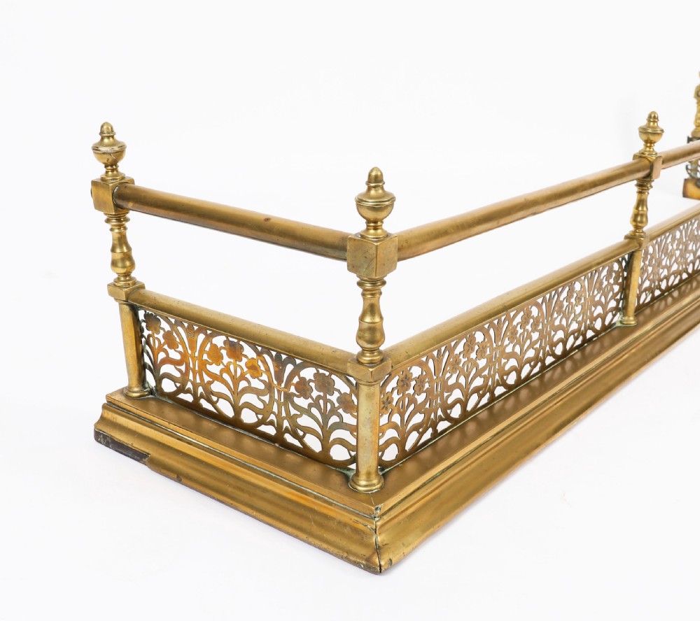 A 19th-century Victorian polished brass fireplace fender featuring a low pierced floral base band topped with a tubular rail supported by finial uprights. View V
