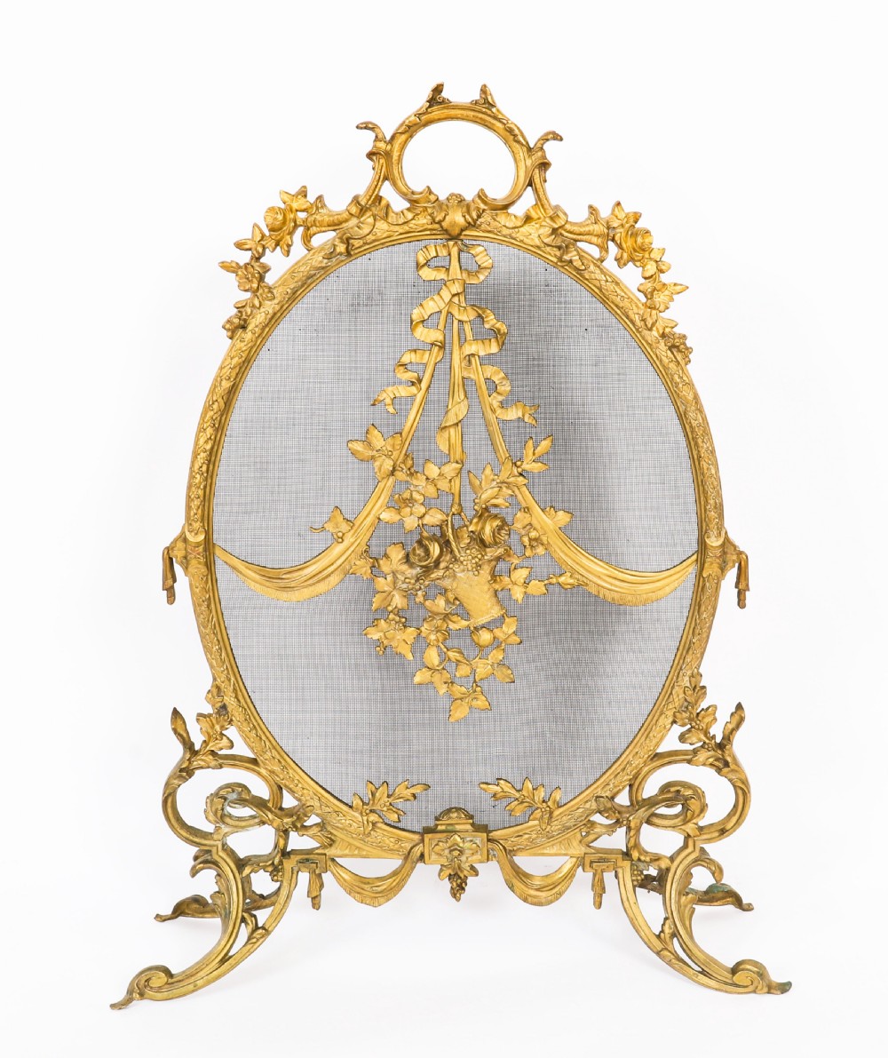 A 19th-century French Rococo Revival brass fire screen with an oval wire mesh insert, ornate bow knot and floral motif, resting on scroll feet. View II