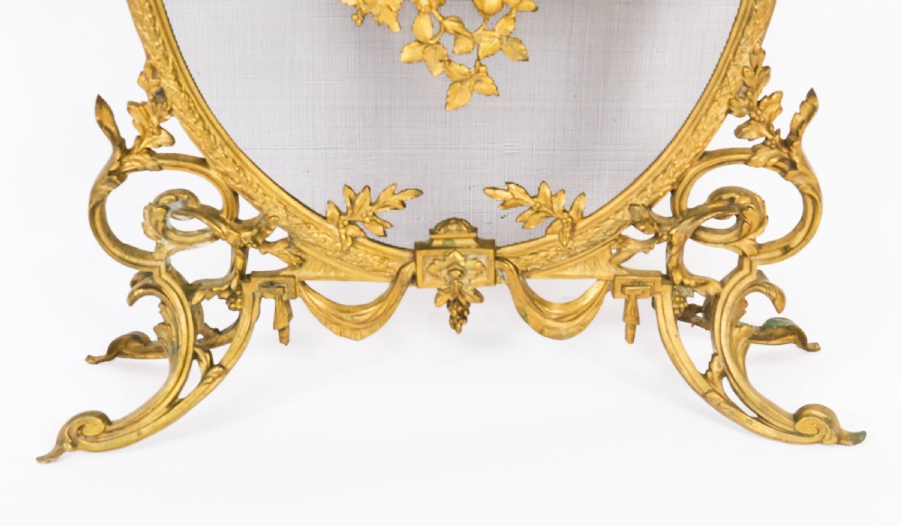 A 19th-century French Rococo Revival brass fire screen with an oval wire mesh insert, ornate bow knot and floral motif, resting on scroll feet. View IV