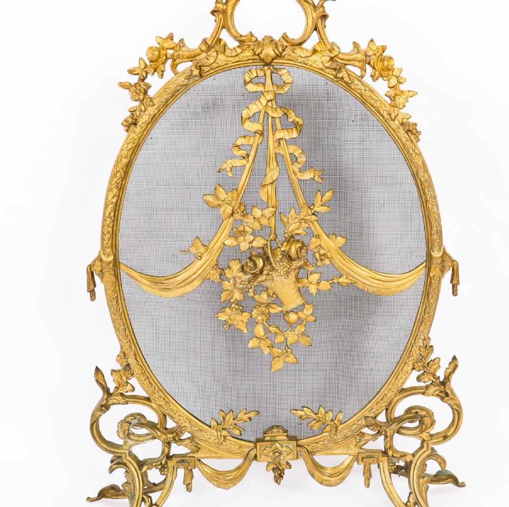 A 19th-century French Rococo Revival brass fire screen with an oval wire mesh insert, ornate bow knot and floral motif, resting on scroll feet. View V