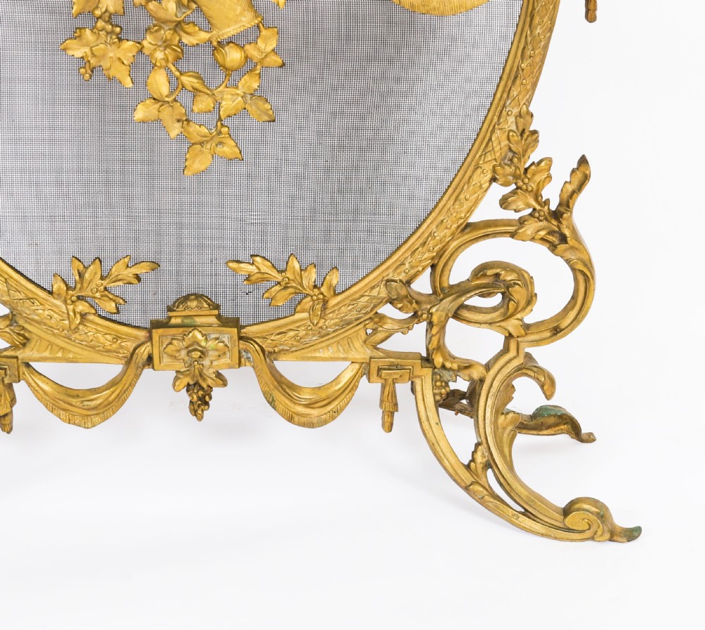 A 19th-century French Rococo Revival brass fire screen with an oval wire mesh insert, ornate bow knot and floral motif, resting on scroll feet. View VI