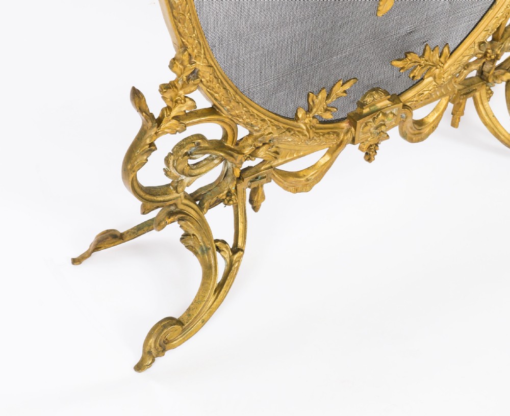 A 19th-century French Rococo Revival brass fire screen with an oval wire mesh insert, ornate bow knot and floral motif, resting on scroll feet. View IX