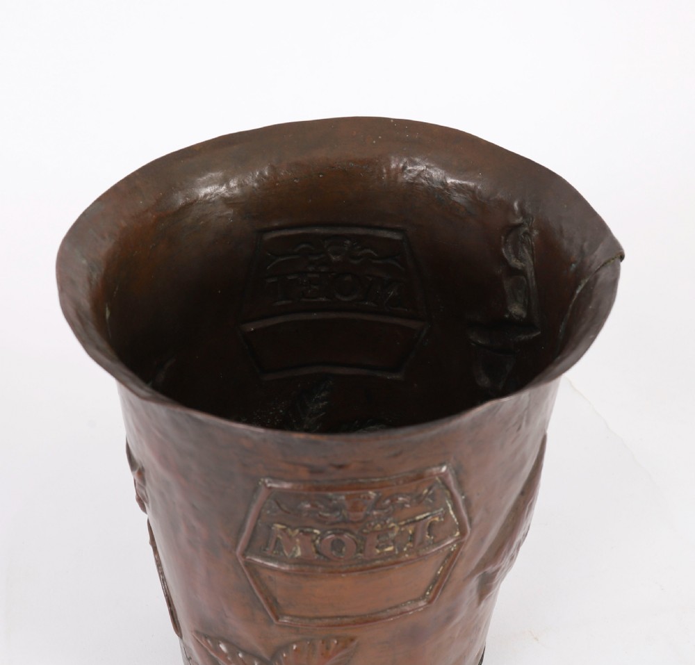 A tapered copper champagne cooler displaying embossed lettering and leaf motifs with a patinated dark surface. View VI