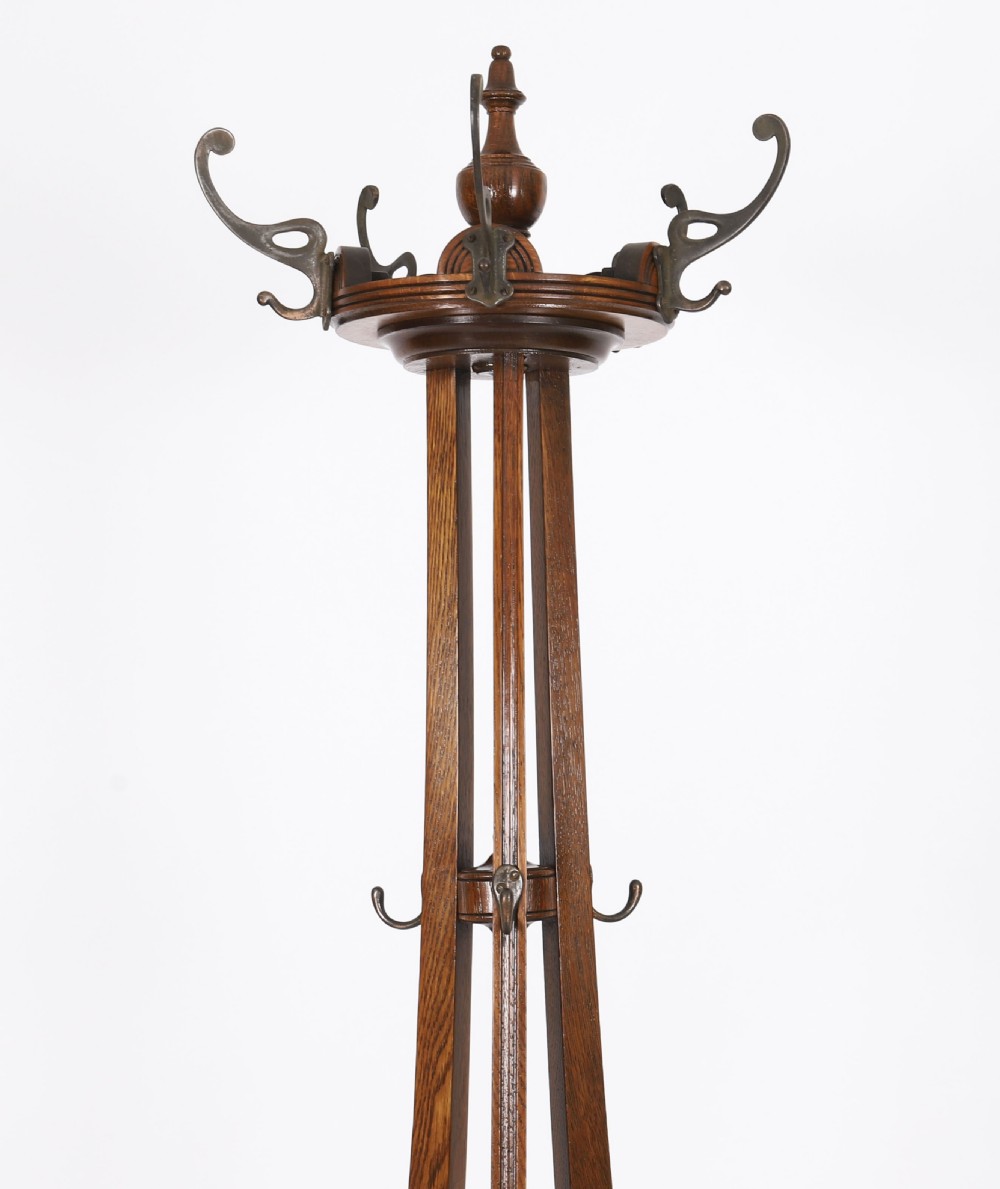 A tall Victorian dark oak coat stand featuring curved supports, upper metal hat hooks, a central brass umbrella retaining ring, and a circular drip tray base. View III
