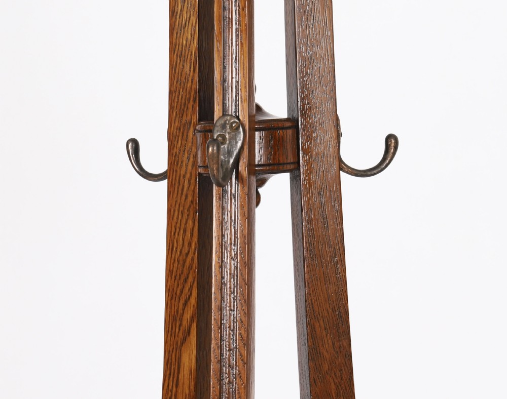 A tall Victorian dark oak coat stand featuring curved supports, upper metal hat hooks, a central brass umbrella retaining ring, and a circular drip tray base. View V