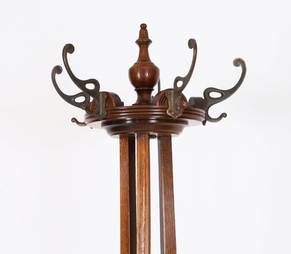 A tall Victorian dark oak coat stand featuring curved supports, upper metal hat hooks, a central brass umbrella retaining ring, and a circular drip tray base. View VI