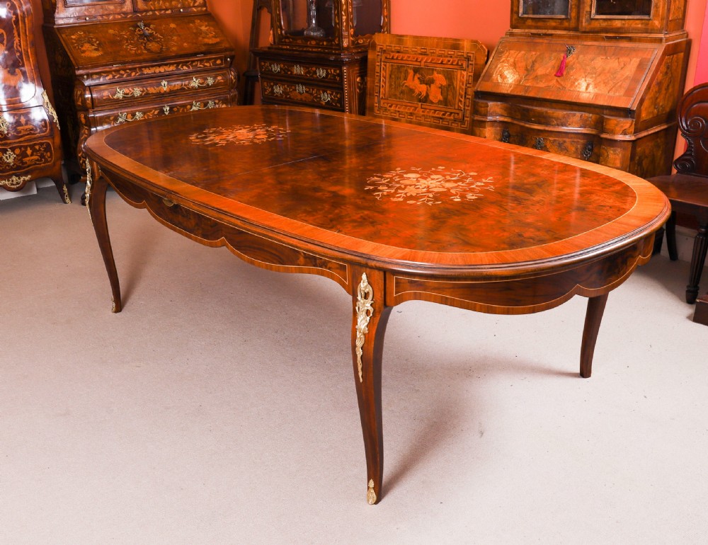 An oval French Louis Revival wooden dining table featuring floral marquetry inlays, serpentine aprons, and gold ormolu leg mounts.