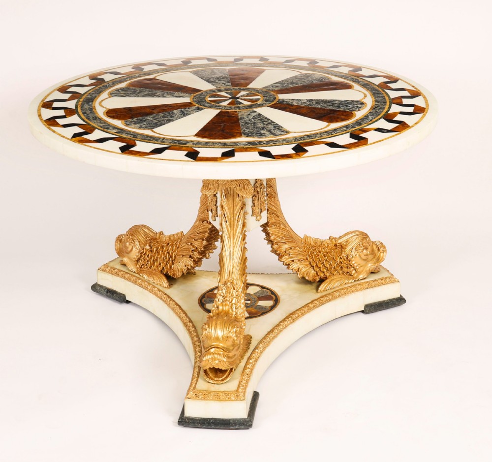 A circular centre table featuring an intricate geometric inlaid marble top supported by three carved gilt dolphin figures on a triform marble base.