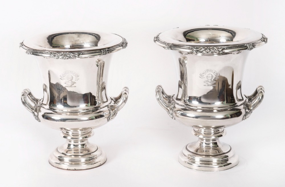 A pair of silver-plated urn-shaped wine coolers featuring scrolled side handles, engraved central crests, and circular pedestal bases.