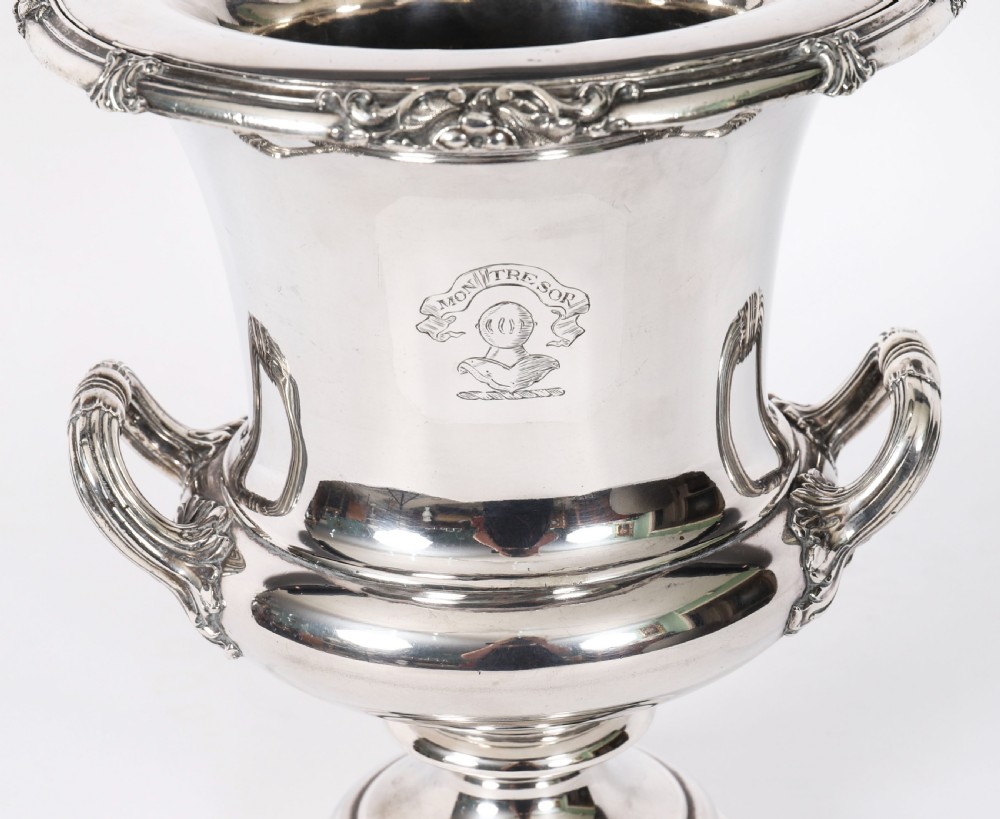 A pair of silver-plated urn-shaped wine coolers featuring scrolled side handles, engraved central crests, and circular pedestal bases. View X