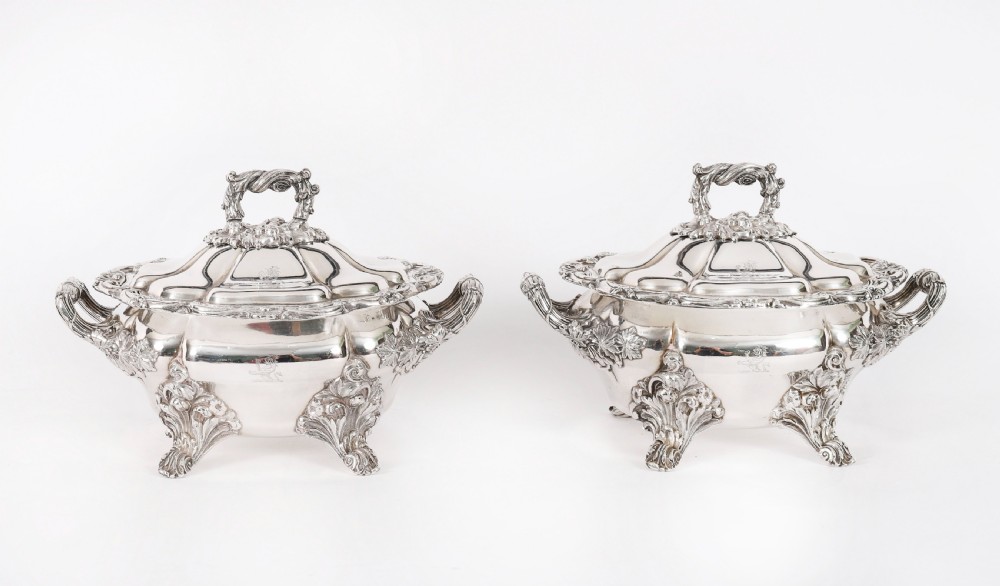A pair of silver-plated oval sauce tureens featuring domed lids, foliate scroll handles, and decorative carved feet.