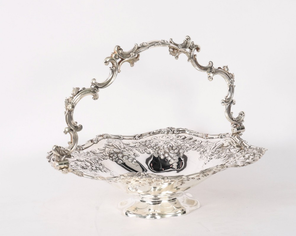 An ornate mid-19th-century Victorian silver-plated fruit basket with a high scroll swing handle, chased leaf patterns, scalloped rim, and pedestal base.