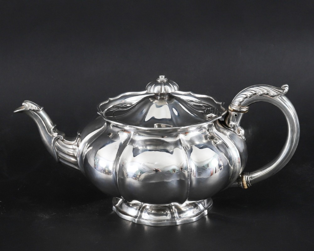 A Georgian sterling silver teapot featuring a lobed melon-shaped body, leaf-engraved spout, and insulated handle. View X