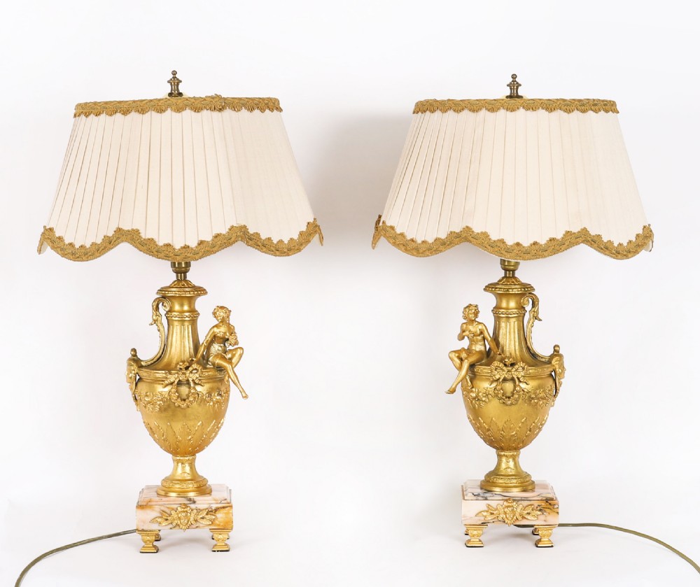 A pair of Neoclassical ormolu urn-form table lamps featuring classical figures mounted on square pink marble bases with cream pleated shades.
