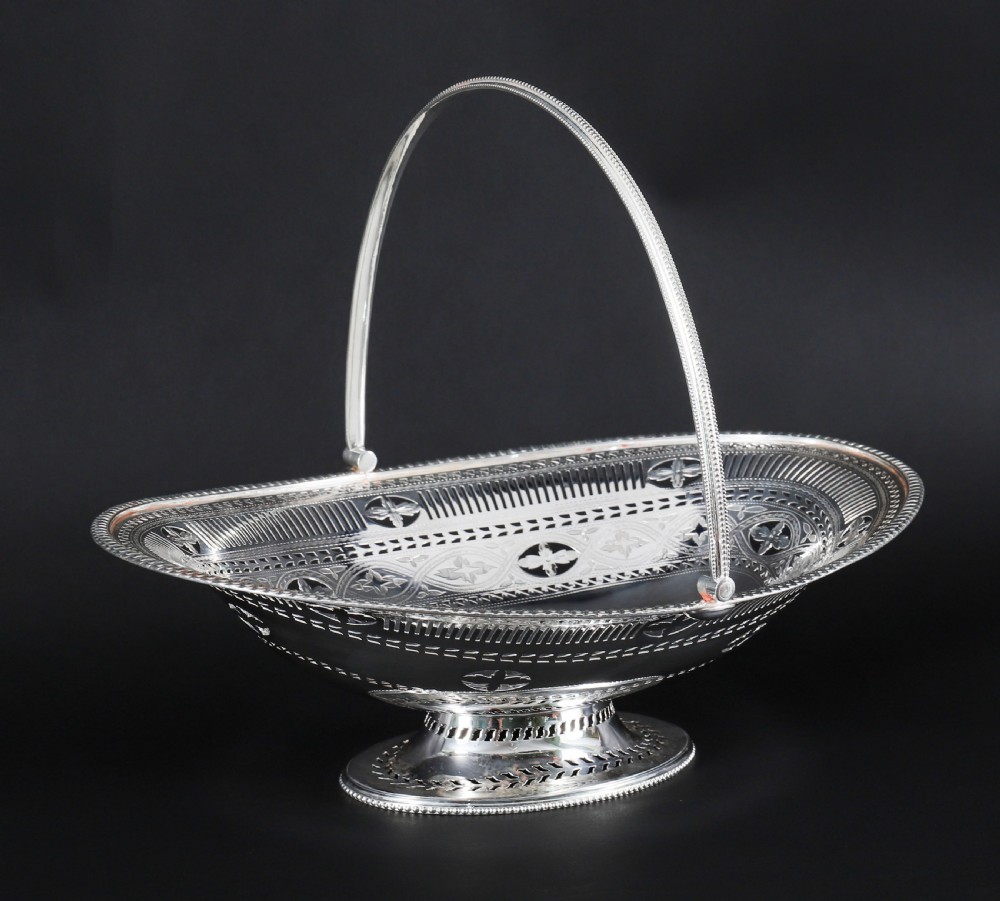 An antique silver-plated oval fruit basket featuring delicate pierced metalwork, engraved geometric motifs, a beaded pedestal foot, and a tall arched handle.