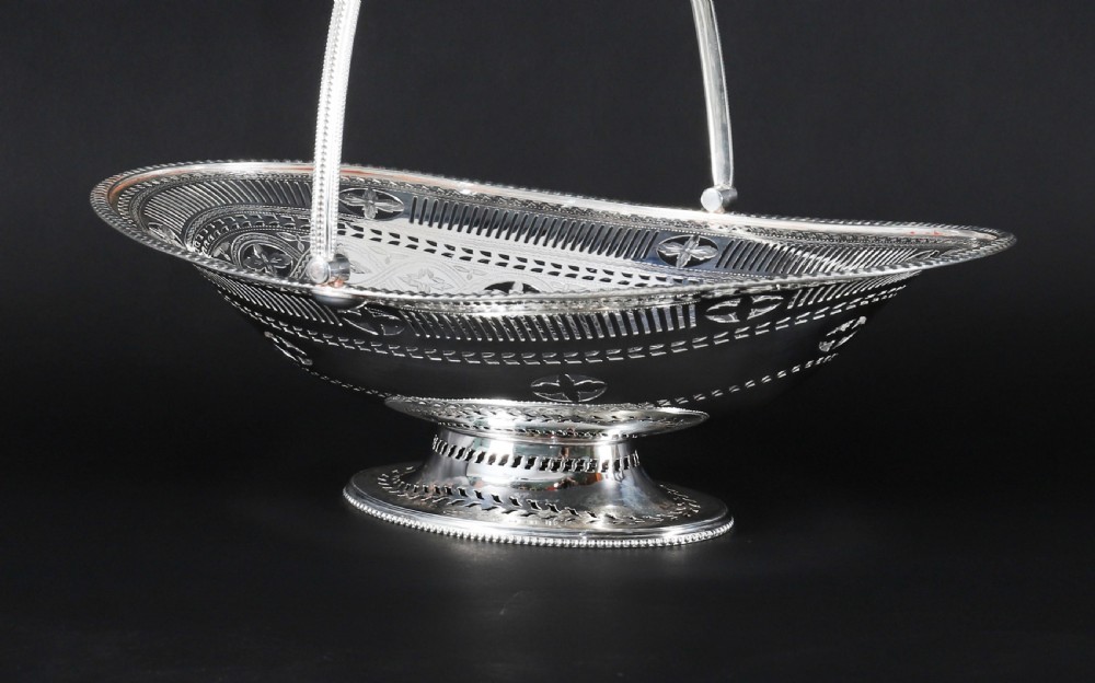 An antique silver-plated oval fruit basket featuring delicate pierced metalwork, engraved geometric motifs, a beaded pedestal foot, and a tall arched handle. View VIII