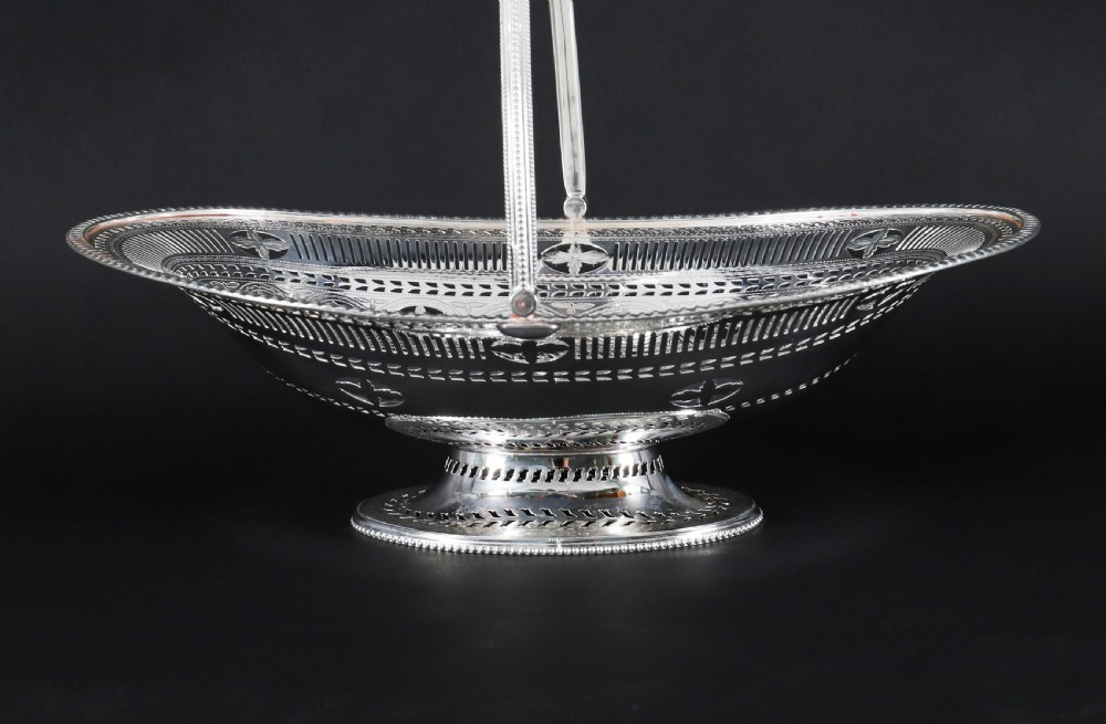 An antique silver-plated oval fruit basket featuring delicate pierced metalwork, engraved geometric motifs, a beaded pedestal foot, and a tall arched handle. View IX