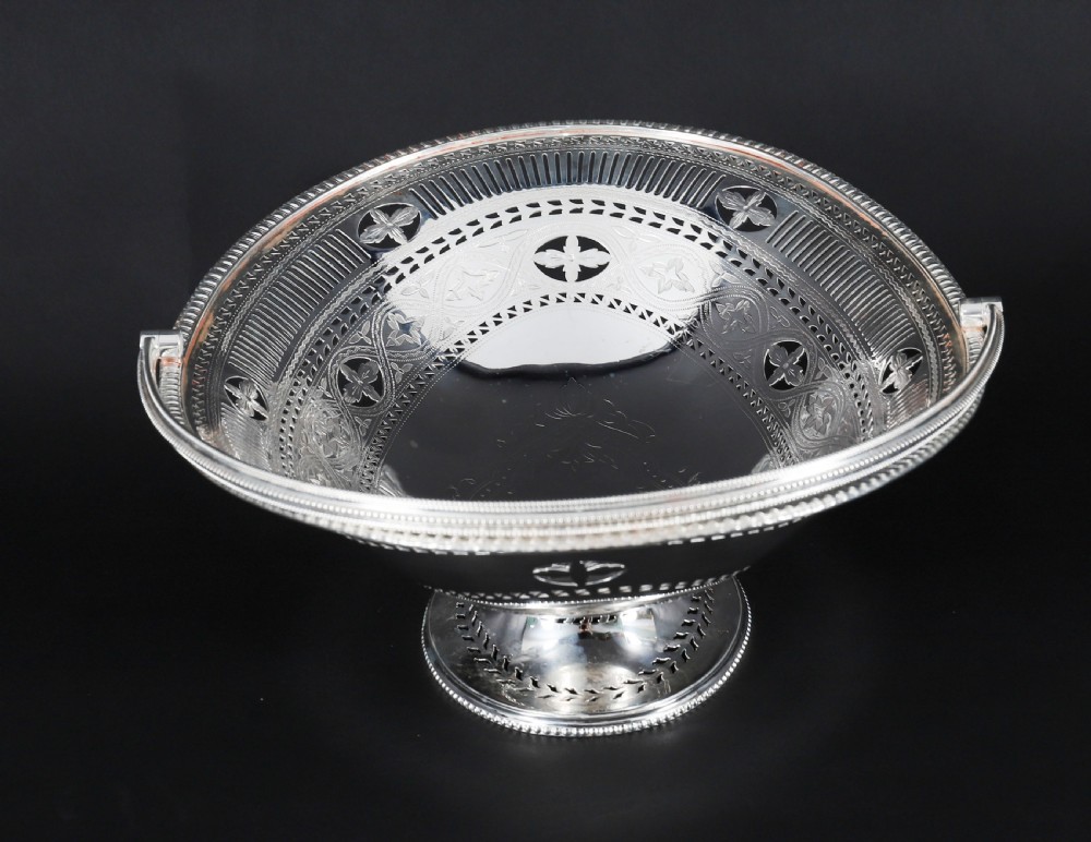 An antique silver-plated oval fruit basket featuring delicate pierced metalwork, engraved geometric motifs, a beaded pedestal foot, and a tall arched handle. View XI