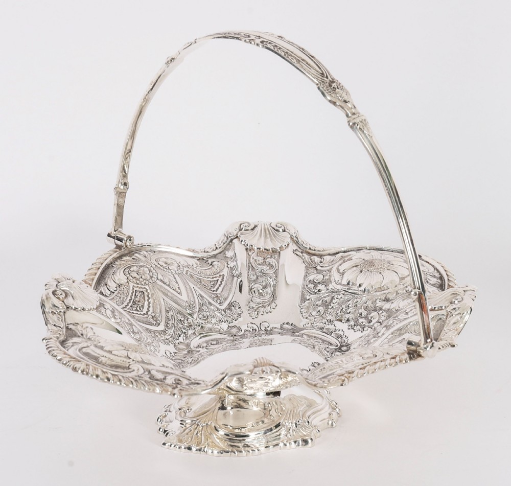 An ornate Victorian silver-plated fruit basket featuring chased floral motifs, a scalloped shell rim, a pedestal base, and a hinged overhead swing handle.