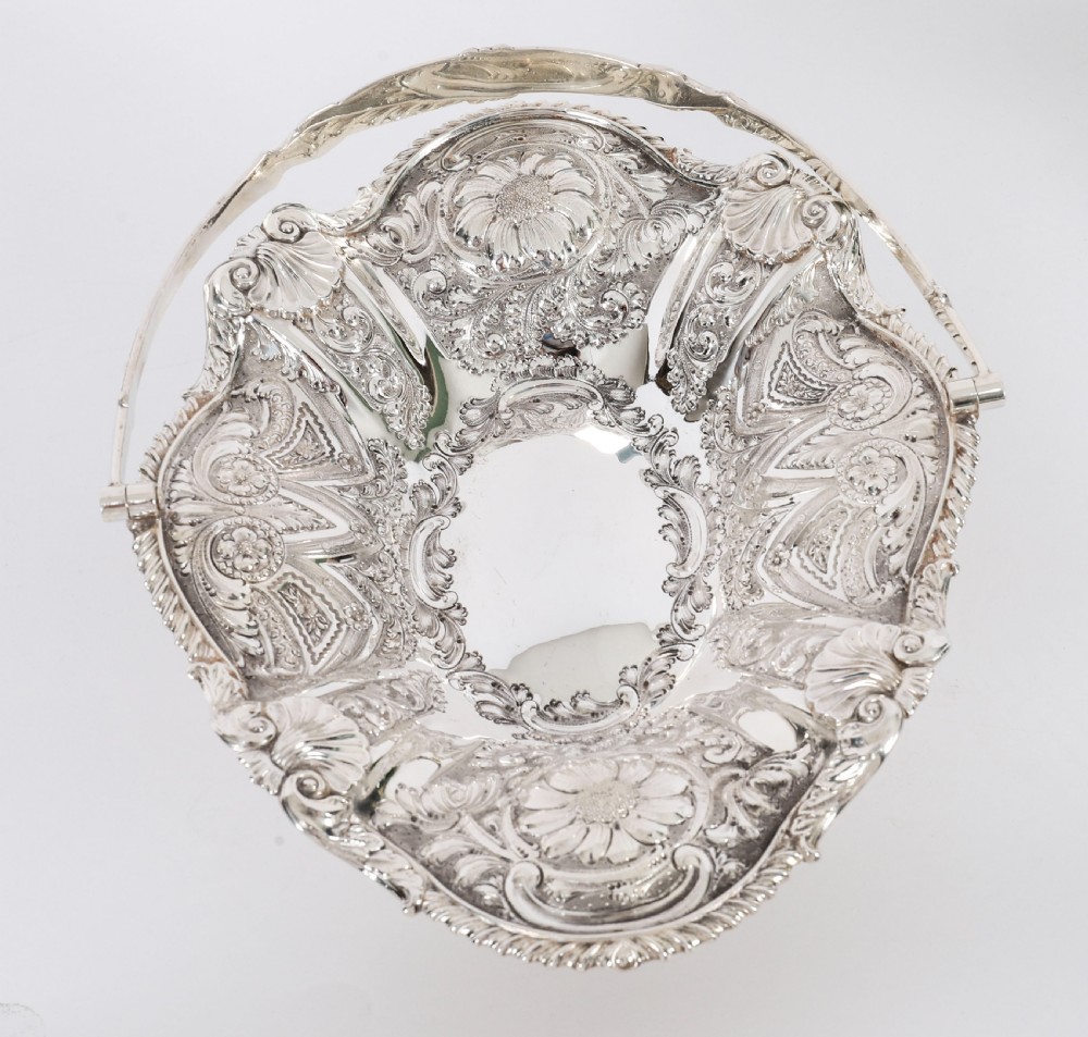 An ornate Victorian silver-plated fruit basket featuring chased floral motifs, a scalloped shell rim, a pedestal base, and a hinged overhead swing handle. View VI
