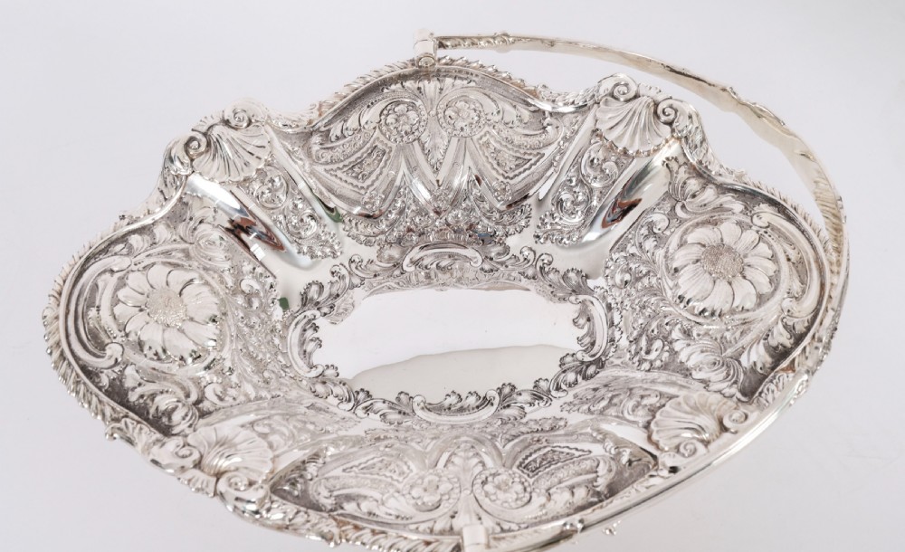 An ornate Victorian silver-plated fruit basket featuring chased floral motifs, a scalloped shell rim, a pedestal base, and a hinged overhead swing handle. View IX