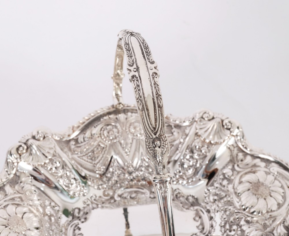 An ornate Victorian silver-plated fruit basket featuring chased floral motifs, a scalloped shell rim, a pedestal base, and a hinged overhead swing handle. View X