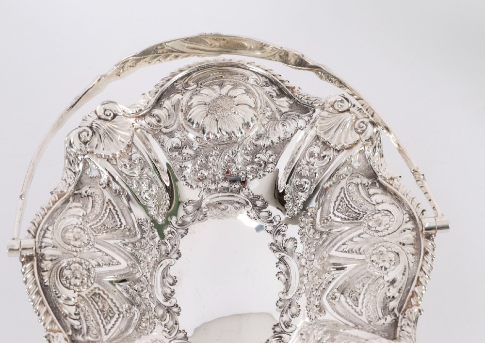 An ornate Victorian silver-plated fruit basket featuring chased floral motifs, a scalloped shell rim, a pedestal base, and a hinged overhead swing handle. View XI