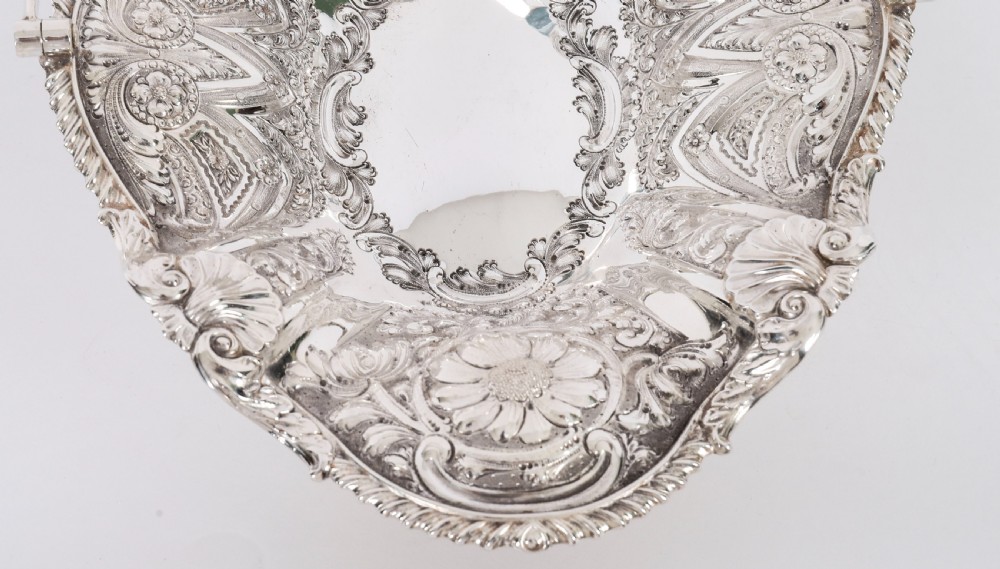 An ornate Victorian silver-plated fruit basket featuring chased floral motifs, a scalloped shell rim, a pedestal base, and a hinged overhead swing handle. View XII