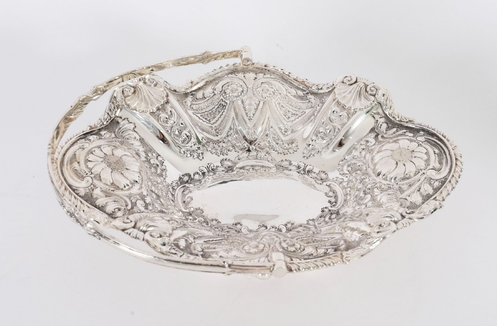 An ornate Victorian silver-plated fruit basket featuring chased floral motifs, a scalloped shell rim, a pedestal base, and a hinged overhead swing handle. View XIII