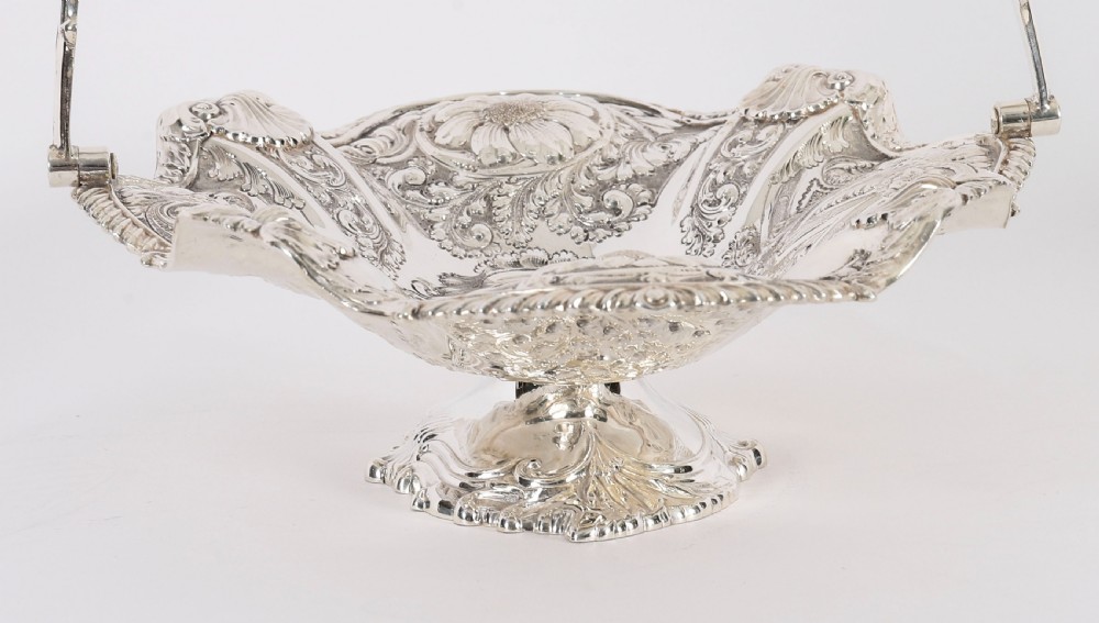 An ornate Victorian silver-plated fruit basket featuring chased floral motifs, a scalloped shell rim, a pedestal base, and a hinged overhead swing handle. View XIV