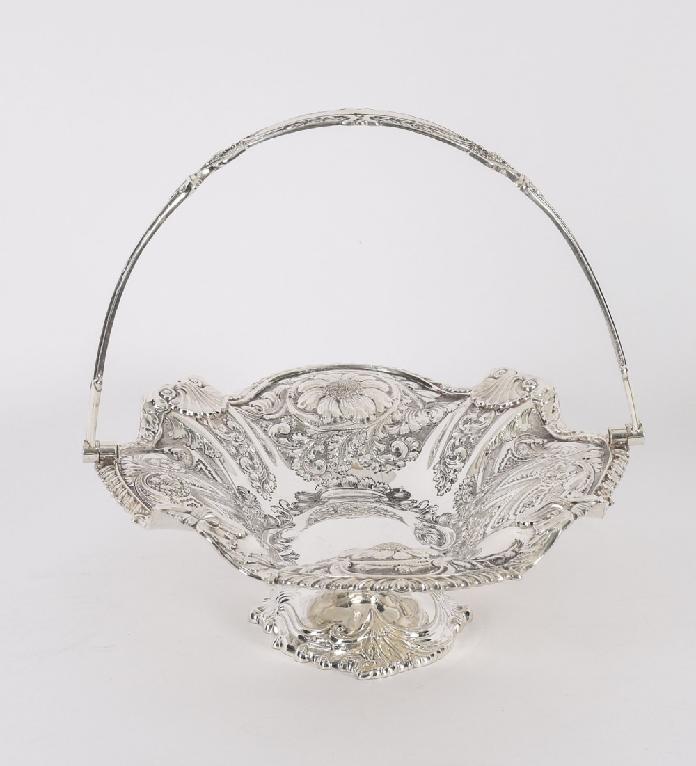 An ornate Victorian silver-plated fruit basket featuring chased floral motifs, a scalloped shell rim, a pedestal base, and a hinged overhead swing handle. View XVI