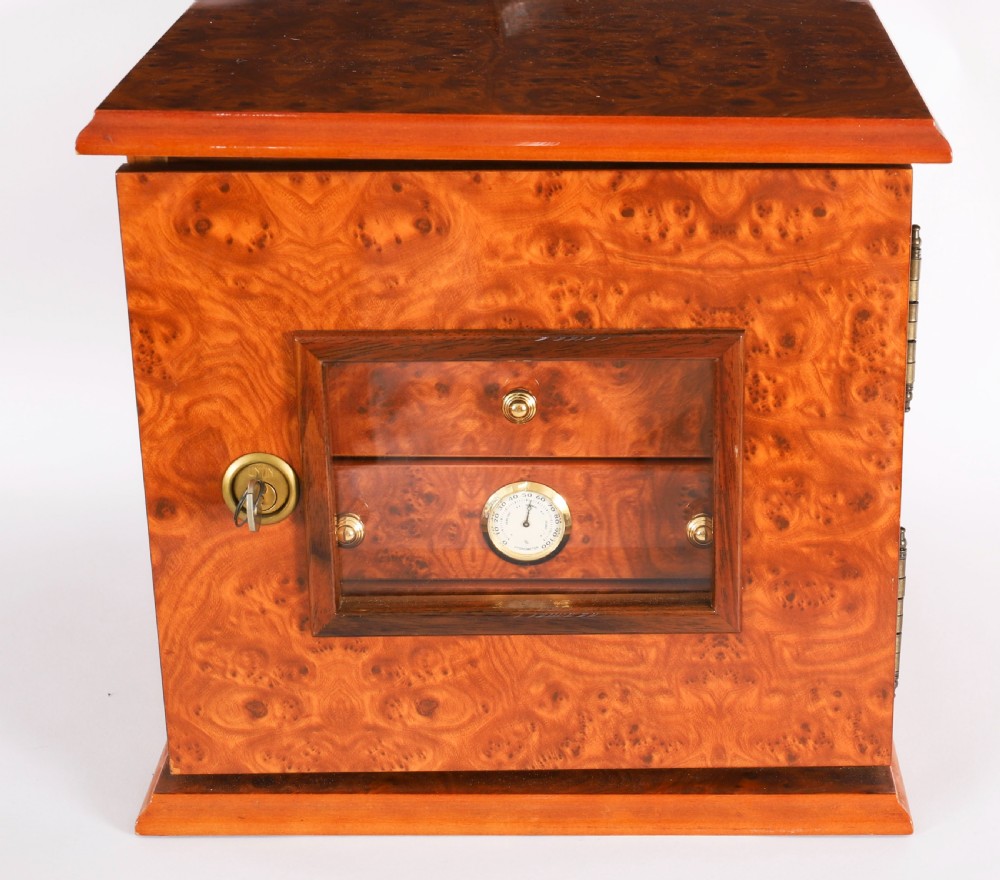 A square burr elm cigar humidor cabinet featuring a glass viewing window, brass hygrometer, front lock, and polished wood figure. View III