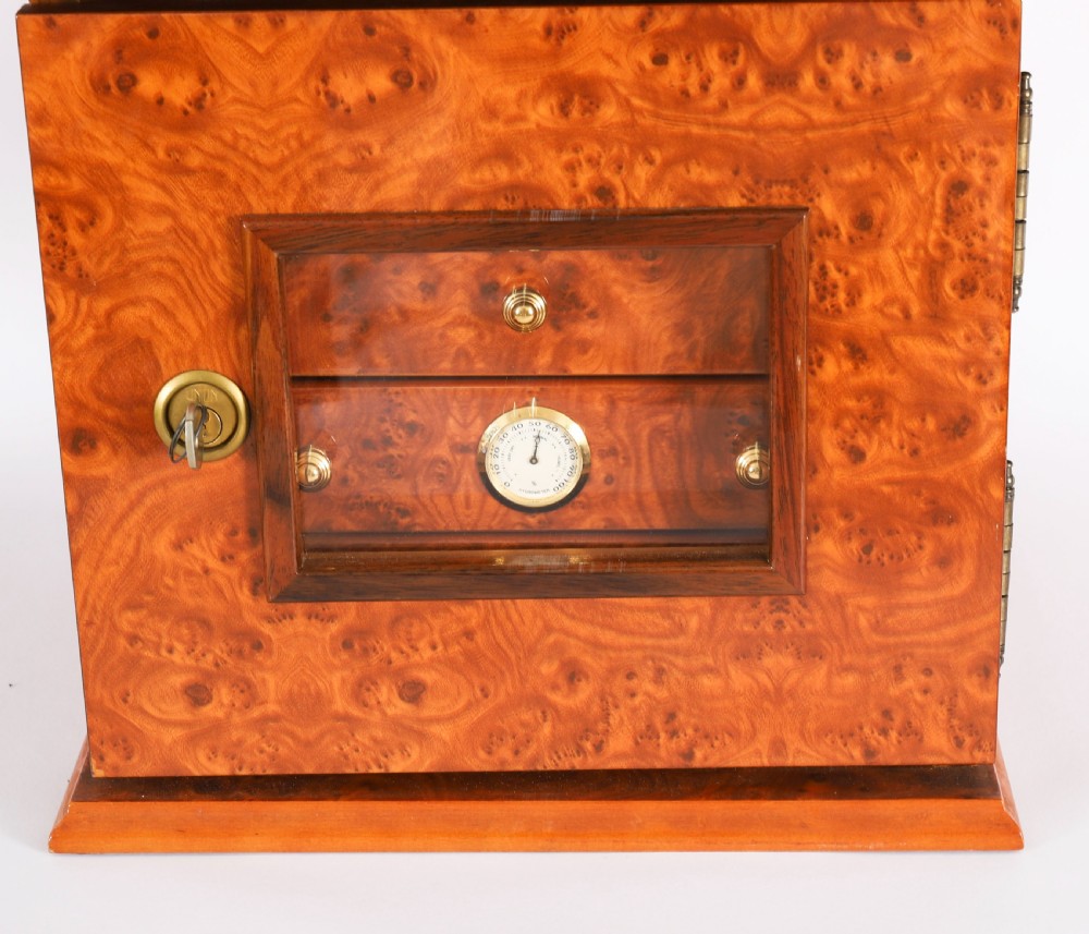 A square burr elm cigar humidor cabinet featuring a glass viewing window, brass hygrometer, front lock, and polished wood figure. View IV