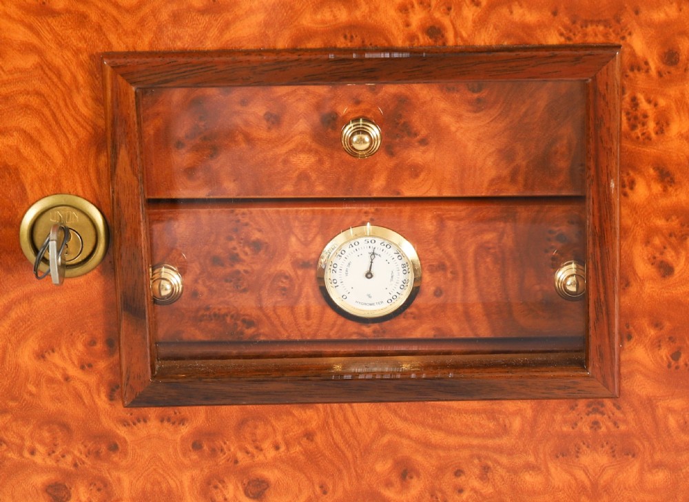 A square burr elm cigar humidor cabinet featuring a glass viewing window, brass hygrometer, front lock, and polished wood figure. View XII
