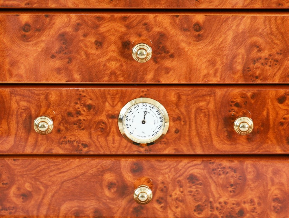 A square burr elm cigar humidor cabinet featuring a glass viewing window, brass hygrometer, front lock, and polished wood figure. View XIII