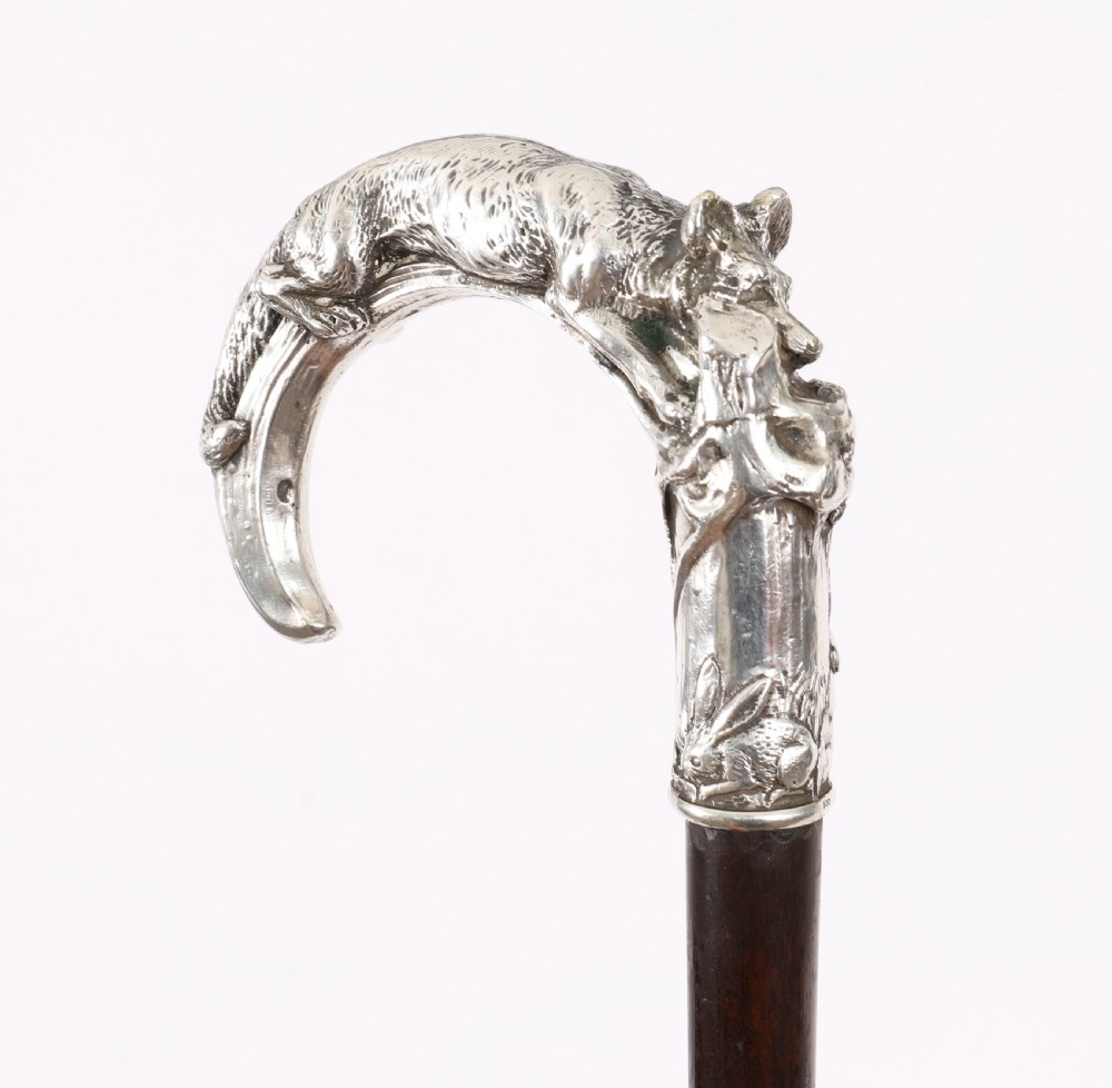 An antique walking stick featuring a detailed silver handle depicting a fox and hare, set atop a dark wood shaft with a metal ferrule. View V