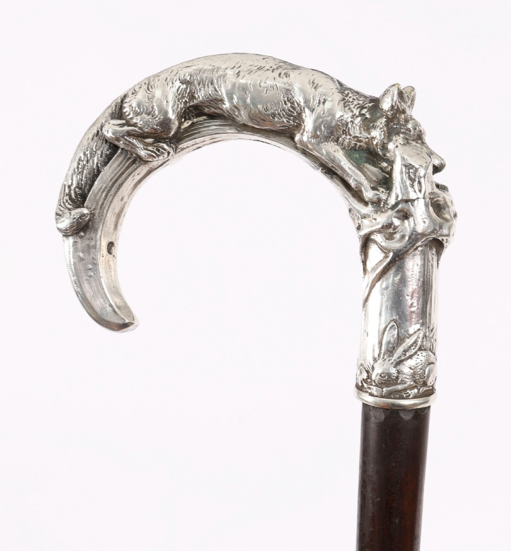 An antique walking stick featuring a detailed silver handle depicting a fox and hare, set atop a dark wood shaft with a metal ferrule. View XVII