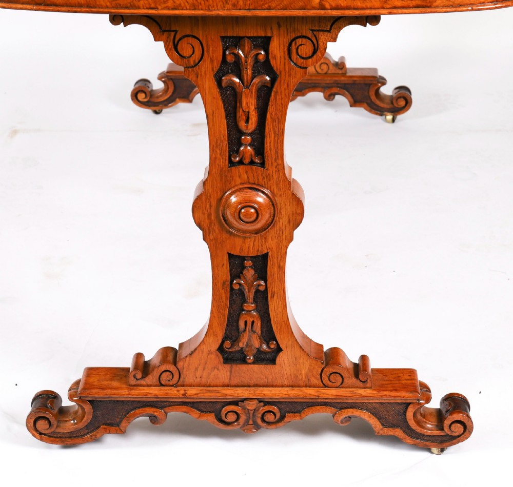 A 19th-century Victorian pollard oak writing table featuring a brown leather inset top, carved trestle end supports, a turned central stretcher, and scroll feet on brass castors. View IX