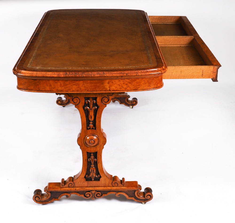 A 19th-century Victorian pollard oak writing table featuring a brown leather inset top, carved trestle end supports, a turned central stretcher, and scroll feet on brass castors. View XII