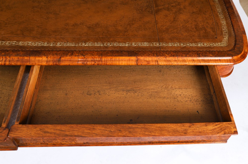 A 19th-century Victorian pollard oak writing table featuring a brown leather inset top, carved trestle end supports, a turned central stretcher, and scroll feet on brass castors. View XIII