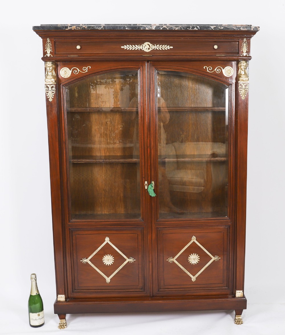 A nineteenth-century French Empire Revival mahogany bookcase featuring twin glazed doors, gilt bronze mounts, a marble top, and metal paw feet. View XIX