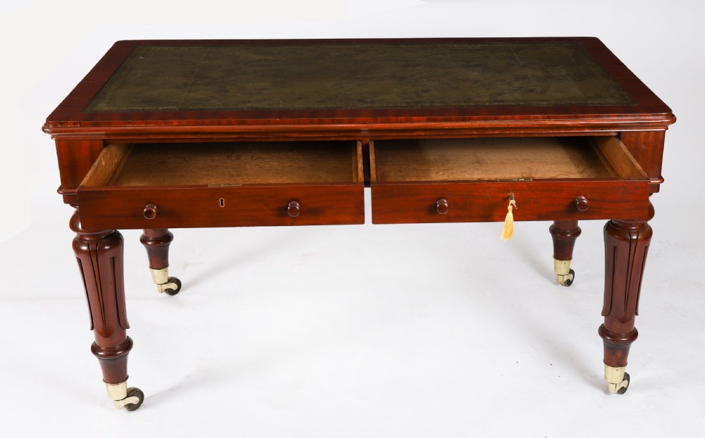 A William IV mahogany writing desk featuring an inset green leather surface, two frieze drawers with knob handles, turned fluted legs, and brass castors. View X