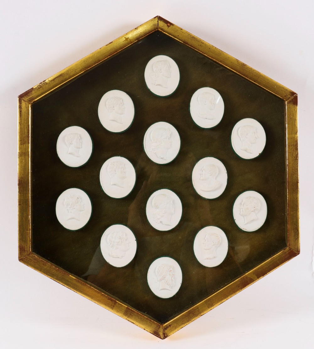 A hexagonal giltwood frame housing thirteen classical white plaster profile intaglios mounted against a dark green background.