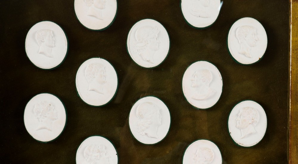 A hexagonal giltwood frame housing thirteen classical white plaster profile intaglios mounted against a dark green background. View V